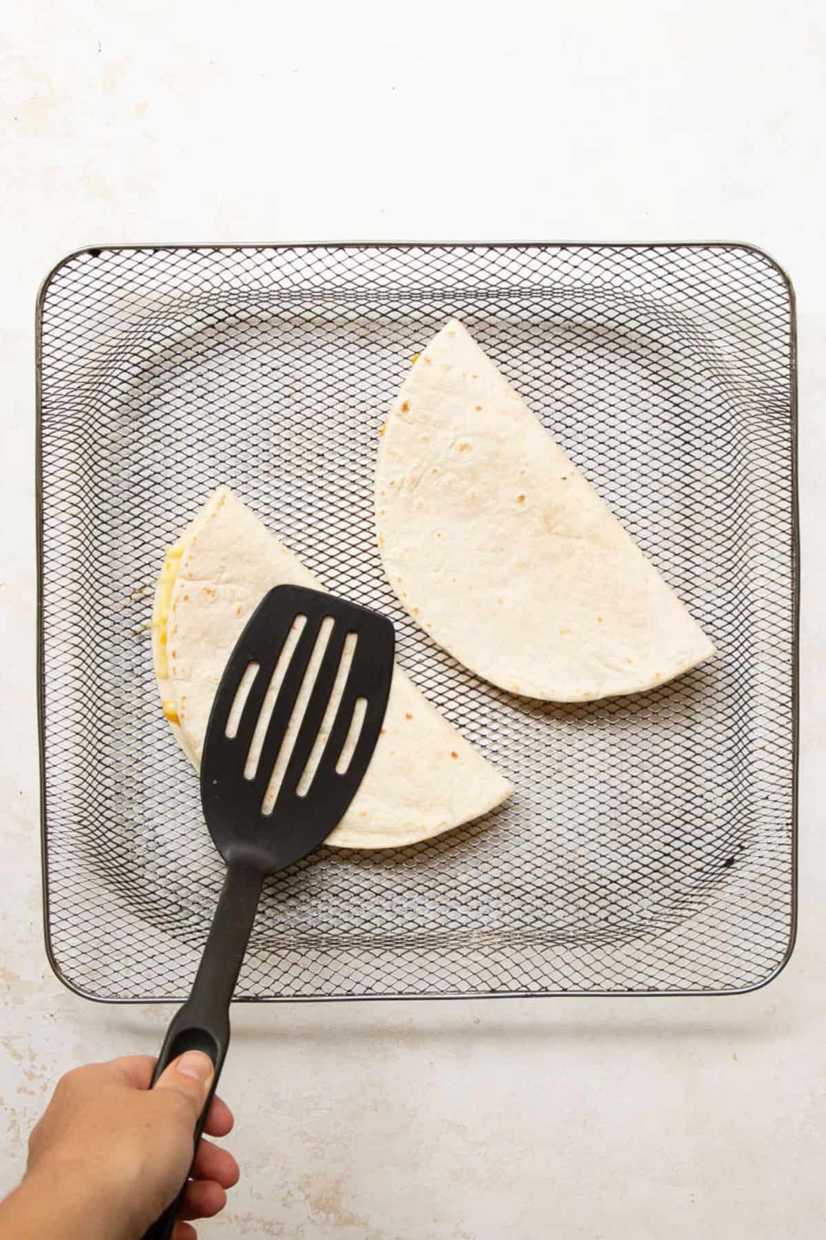 Pressing a folded quesadilla flat with a black spatula in the air fryer basket.