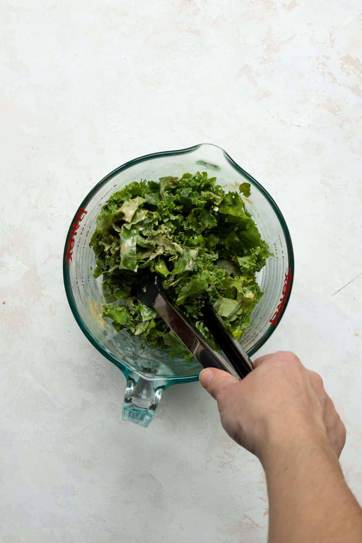 Massaging chopped kale with tongs in a glass mixing bowl.