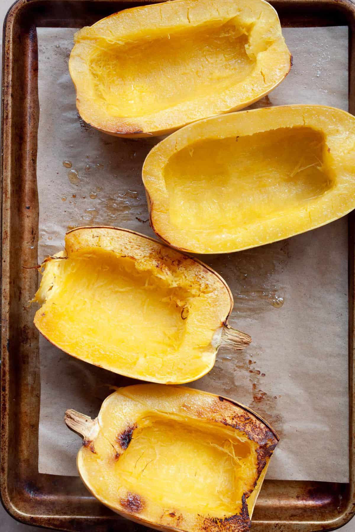 Four roasted spaghetti squash halves with browned edges on a parchment-lined baking sheet.