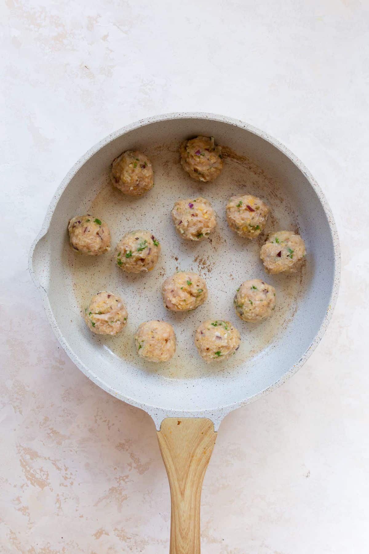 Raw Greek chicken meatballs spaced evenly in an oiled white skillet before searing.