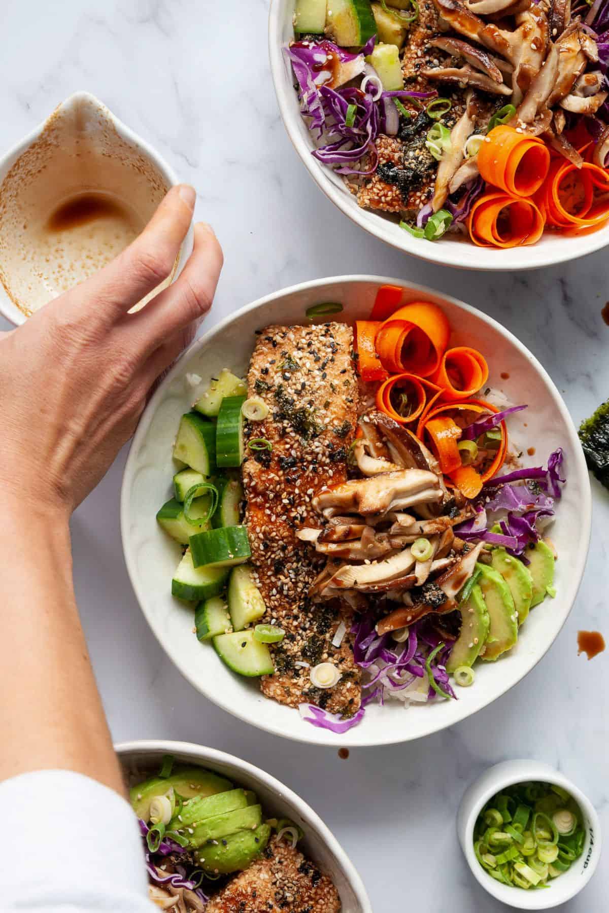Hand pouring honey soy dressing over a furikake salmon bowl with cucumber, carrots, and shiitake mushrooms.