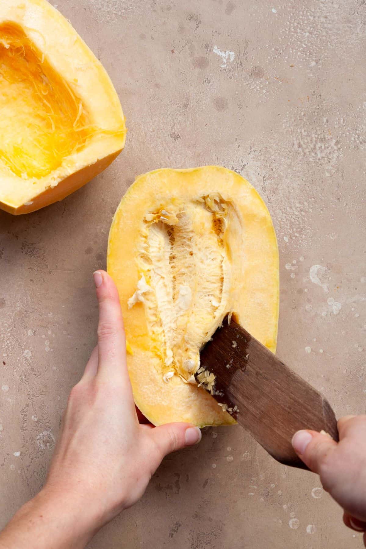 Hands scraping the seeds out of a halved raw spaghetti squash with a wooden spoon.