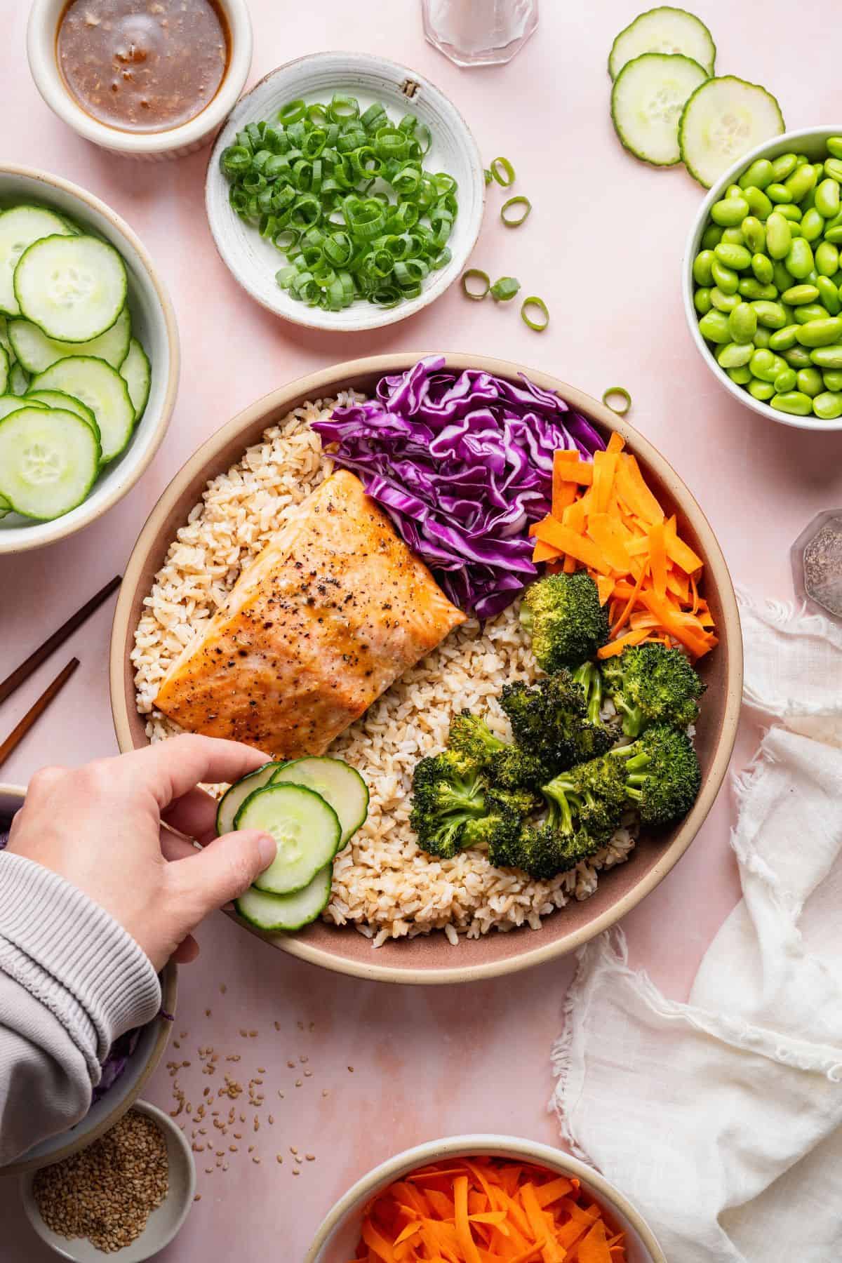 Adding sliced cucumber to a salmon bowl with brown rice, roasted broccoli, red cabbage, and carrots.
