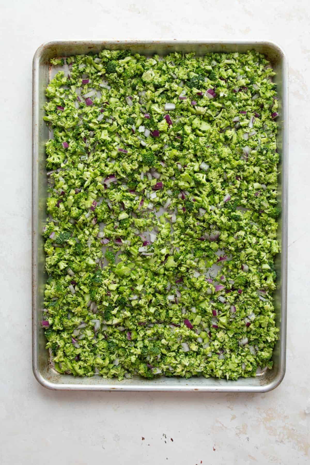 Chopped broccoli and red onion spread in a single layer on a sheet pan before roasting.