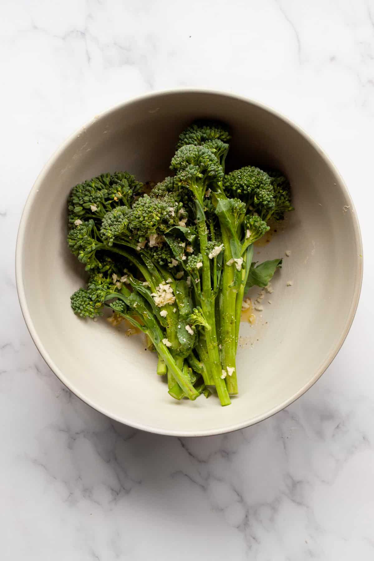 Broccolini tossed with olive oil, minced garlic, salt, and pepper in a white mixing bowl.