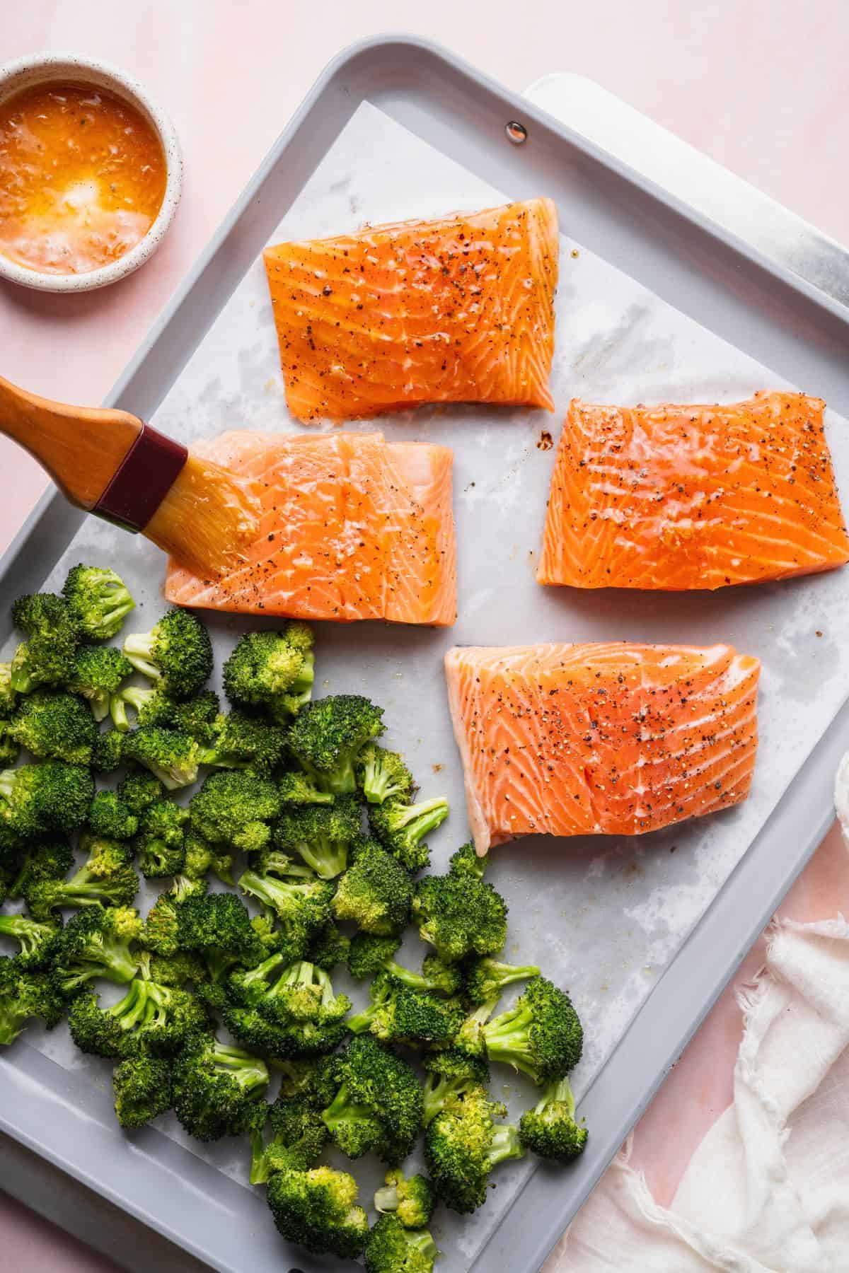 Brushing homemade teriyaki glaze onto salmon fillets next to roasted broccoli on a sheet pan.