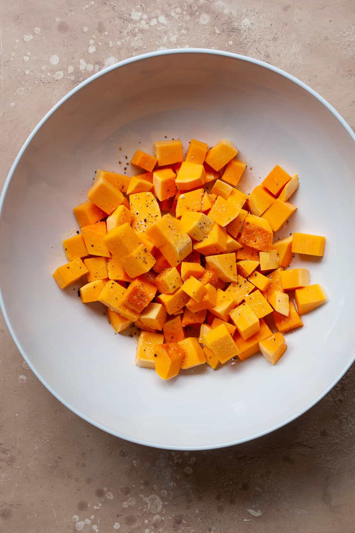 Raw butternut squash cubes in a white bowl with olive oil, salt, and black pepper.