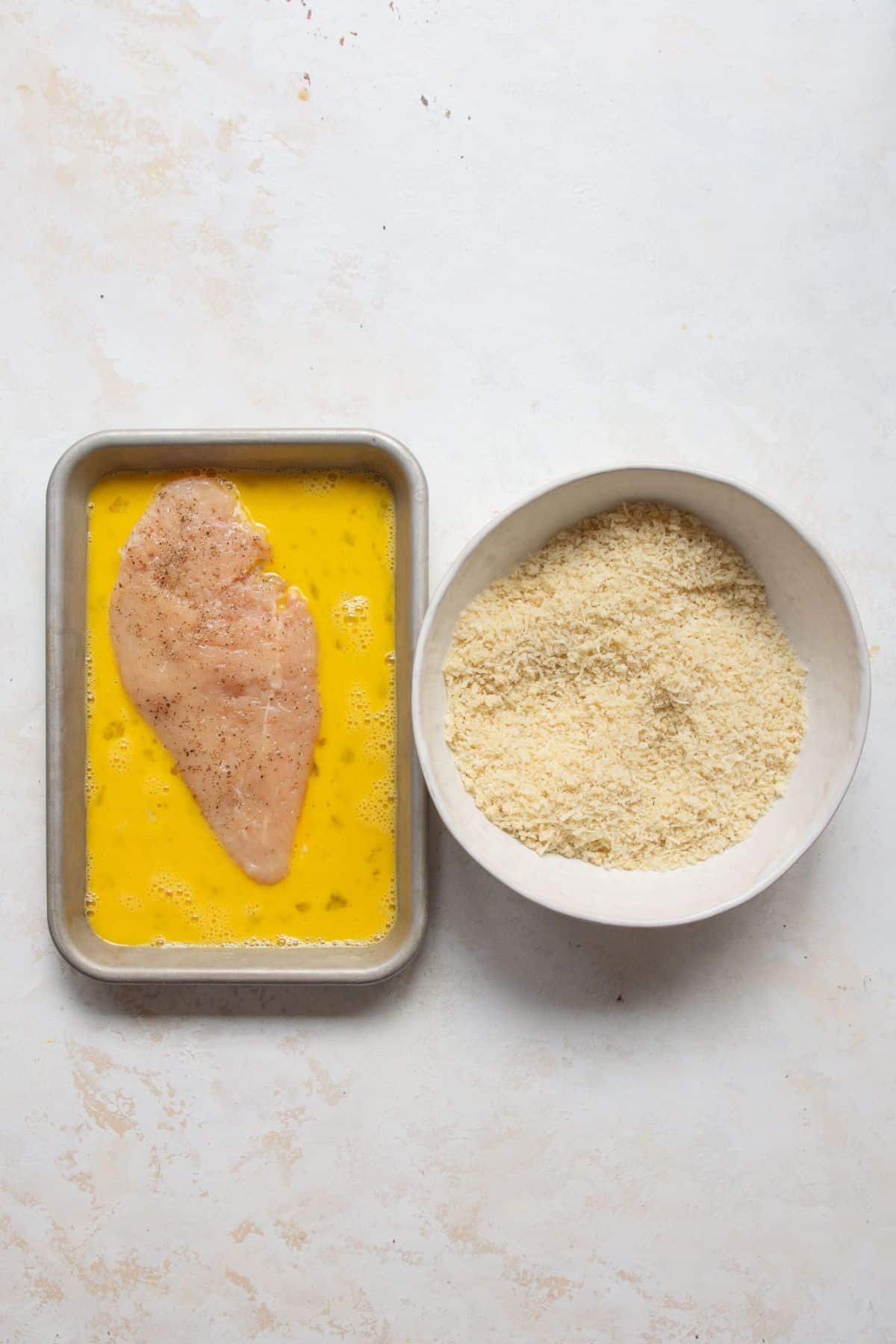 Dipping a seasoned chicken cutlet in beaten eggs, next to a bowl of panko and Parmesan.