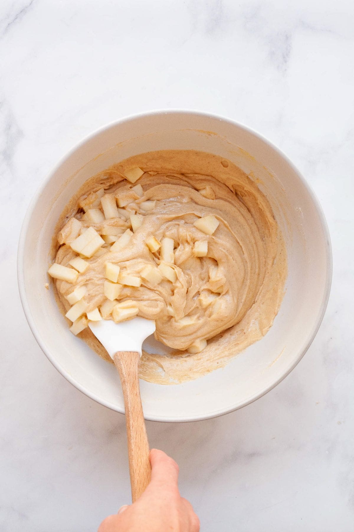 Chopped apples being folded into gluten-free cake batter with a spatula.
