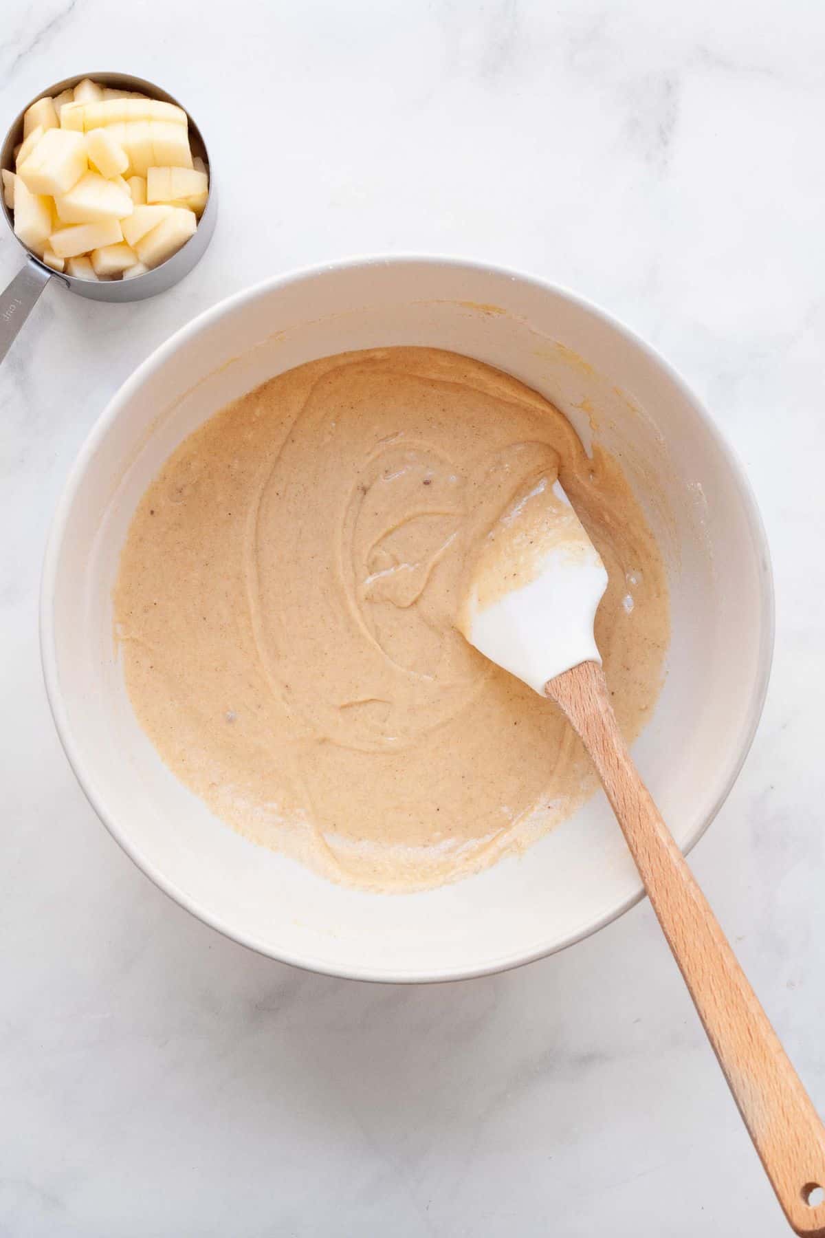 Thick gluten-free apple cake batter being stirred with a wooden spatula in a white bowl.