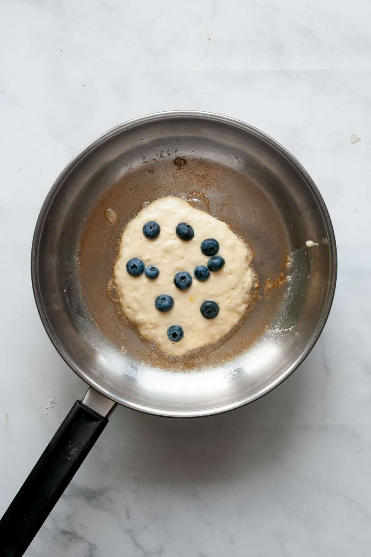 Pancake batter cooking in a buttered skillet with fresh blueberries arranged on top.
