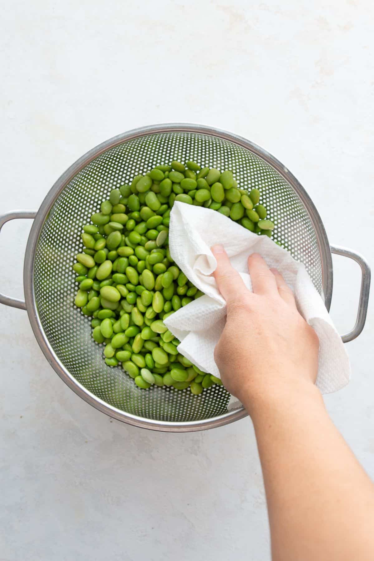 Patting thawed shelled edamame dry with a towel in a colander.