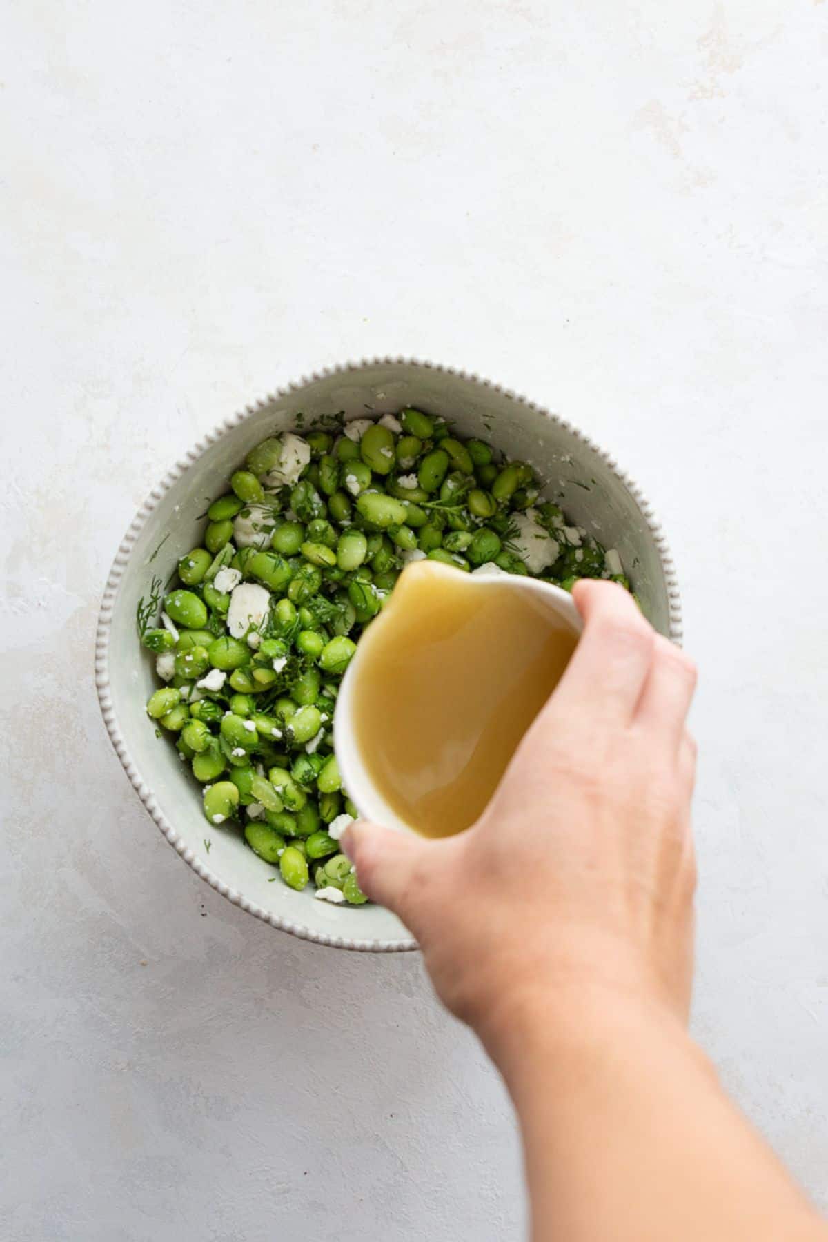 Pouring apple cider vinegar dressing over edamame, feta, and dill in a mixing bowl.