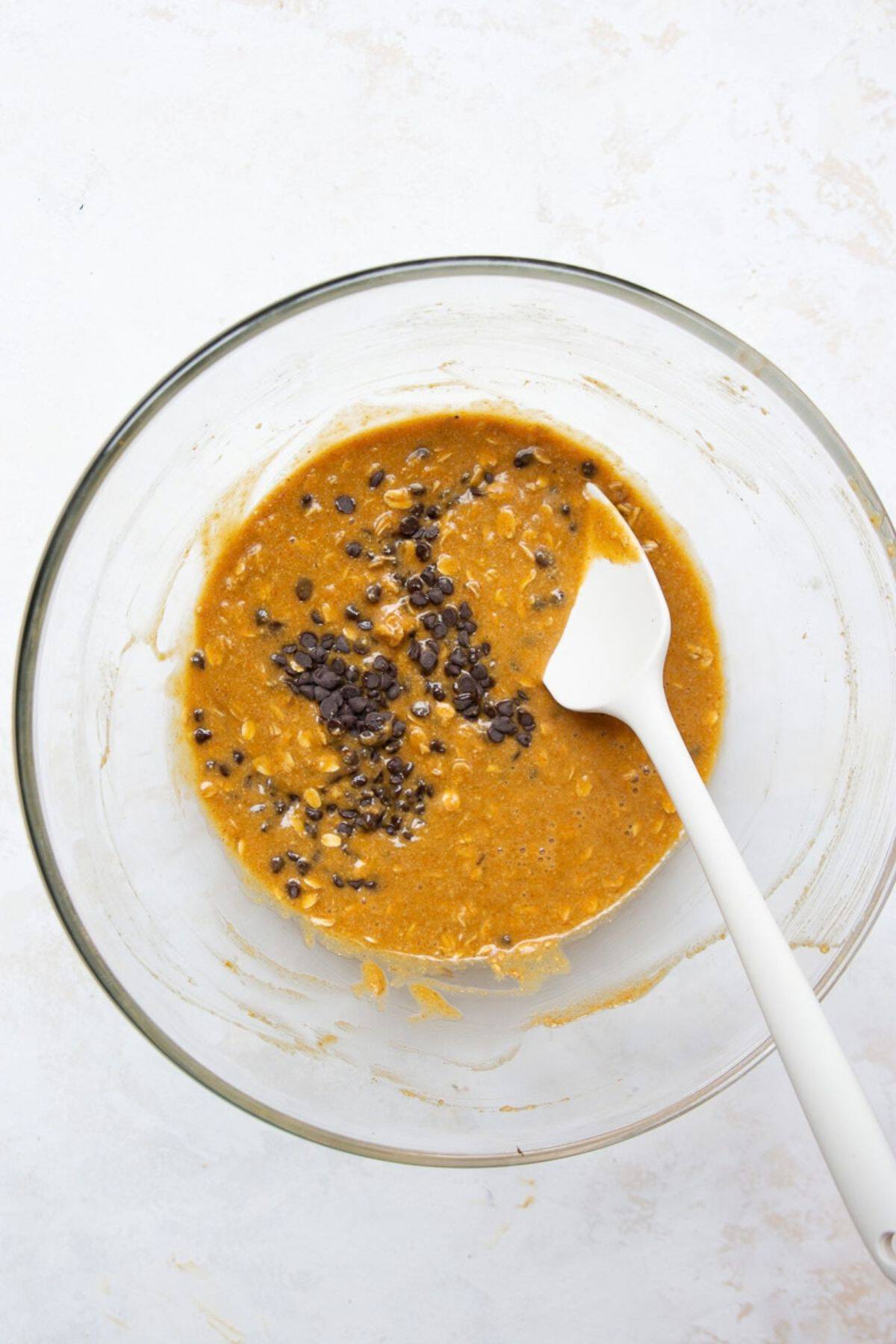Mini chocolate chips added to pumpkin oat batter in a glass mixing bowl.