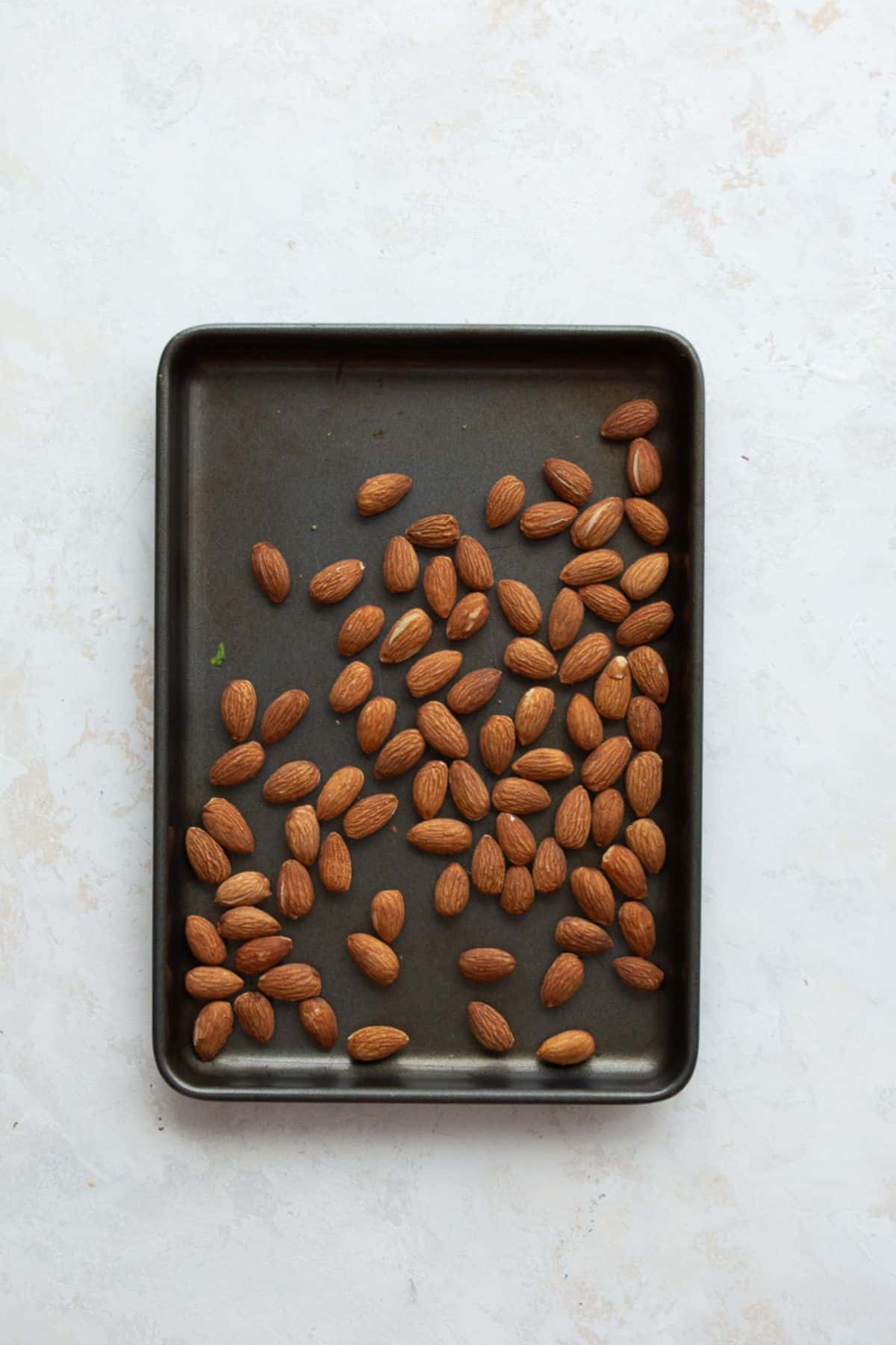 Raw almonds spread on a small dark sheet pan for toasting.