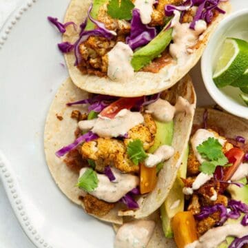 Roasted cauliflower tacos in corn tortillas with refried beans, avocado slices, tomato, red cabbage, cilantro, and homemade yogurt crema.