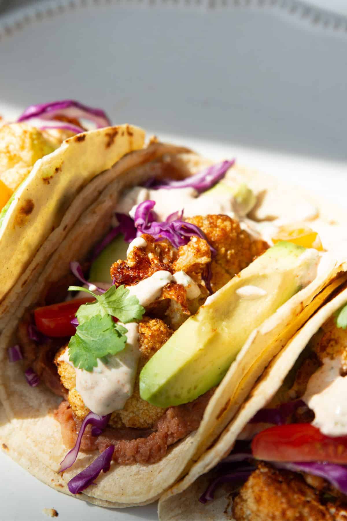Close-up of a crispy cauliflower taco with refried beans, roasted cauliflower, avocado, cherry tomatoes, red cabbage, and yogurt crema.