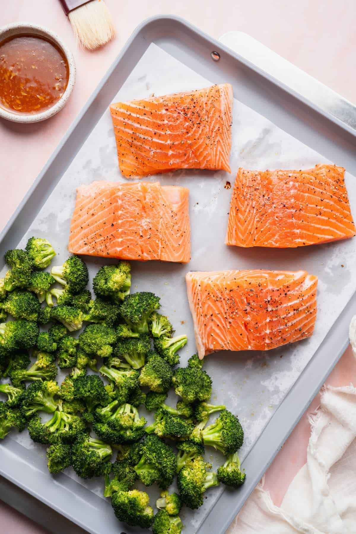 Four salmon fillets seasoned with salt and pepper next to roasted broccoli on a sheet pan.