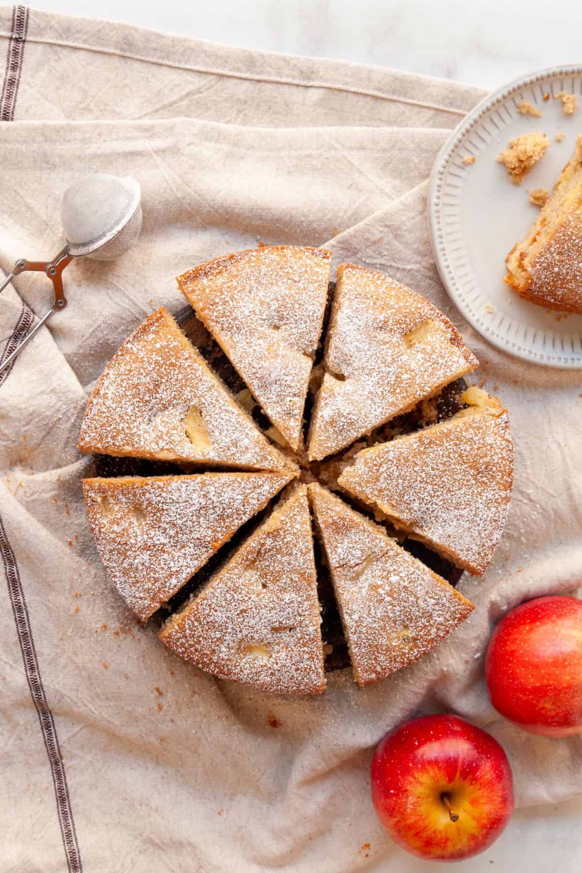 Gluten-free apple snack cake cut into wedges and dusted with powdered sugar, with red apples on the side.