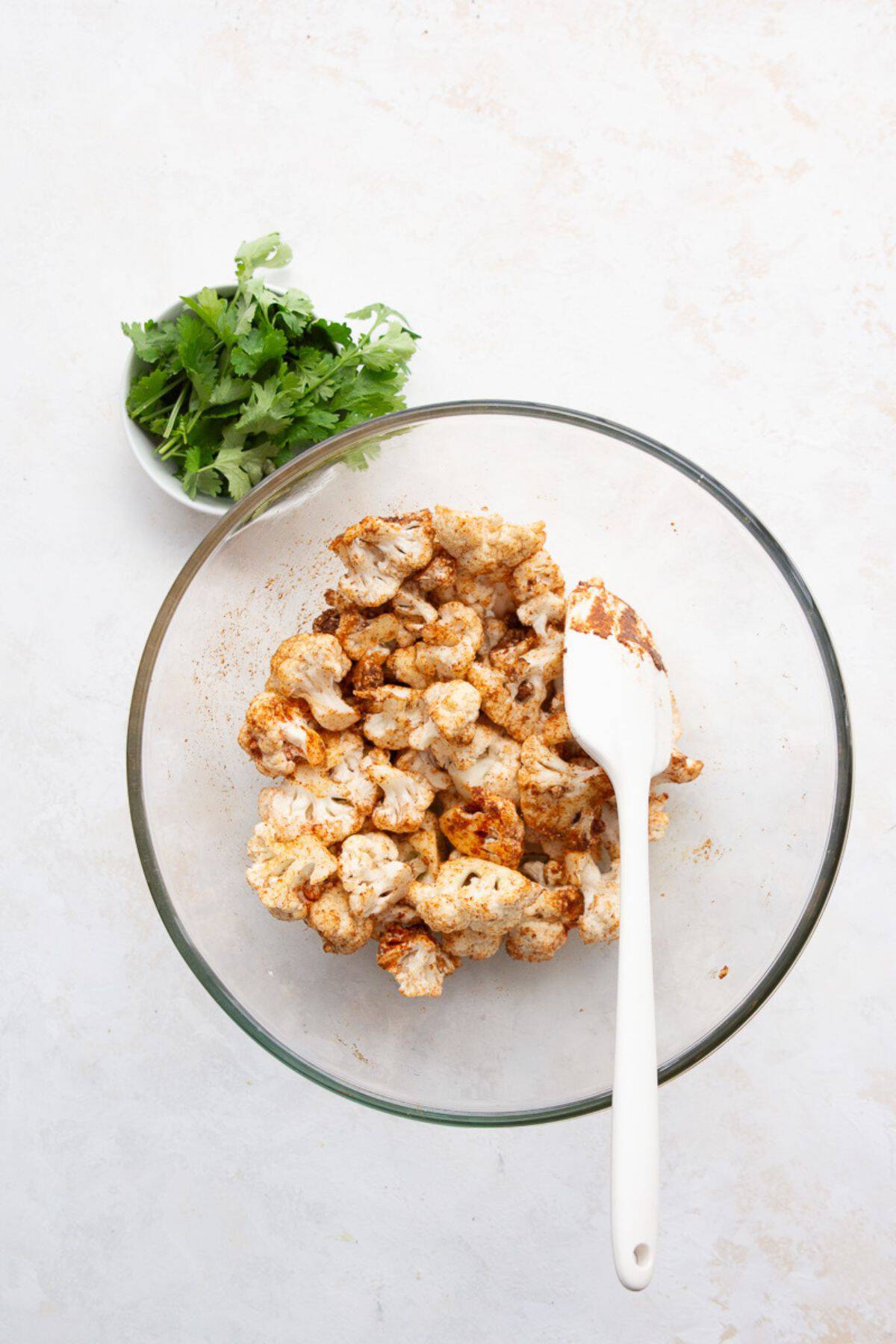 Raw cauliflower florets tossed with oil and spices in a glass mixing bowl with a white spatula.
