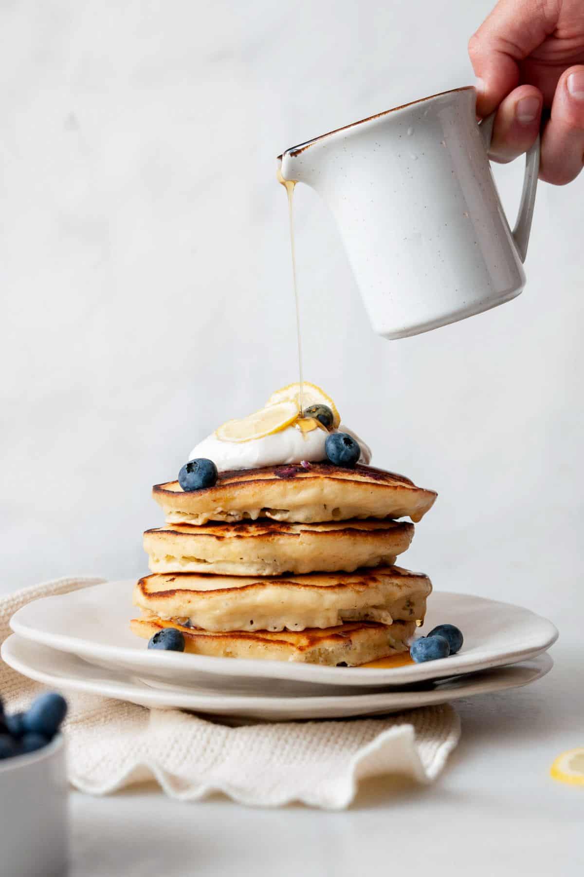 Maple syrup being poured over a stack of lemon blueberry pancakes topped with Greek yogurt, lemon slices, and blueberries.