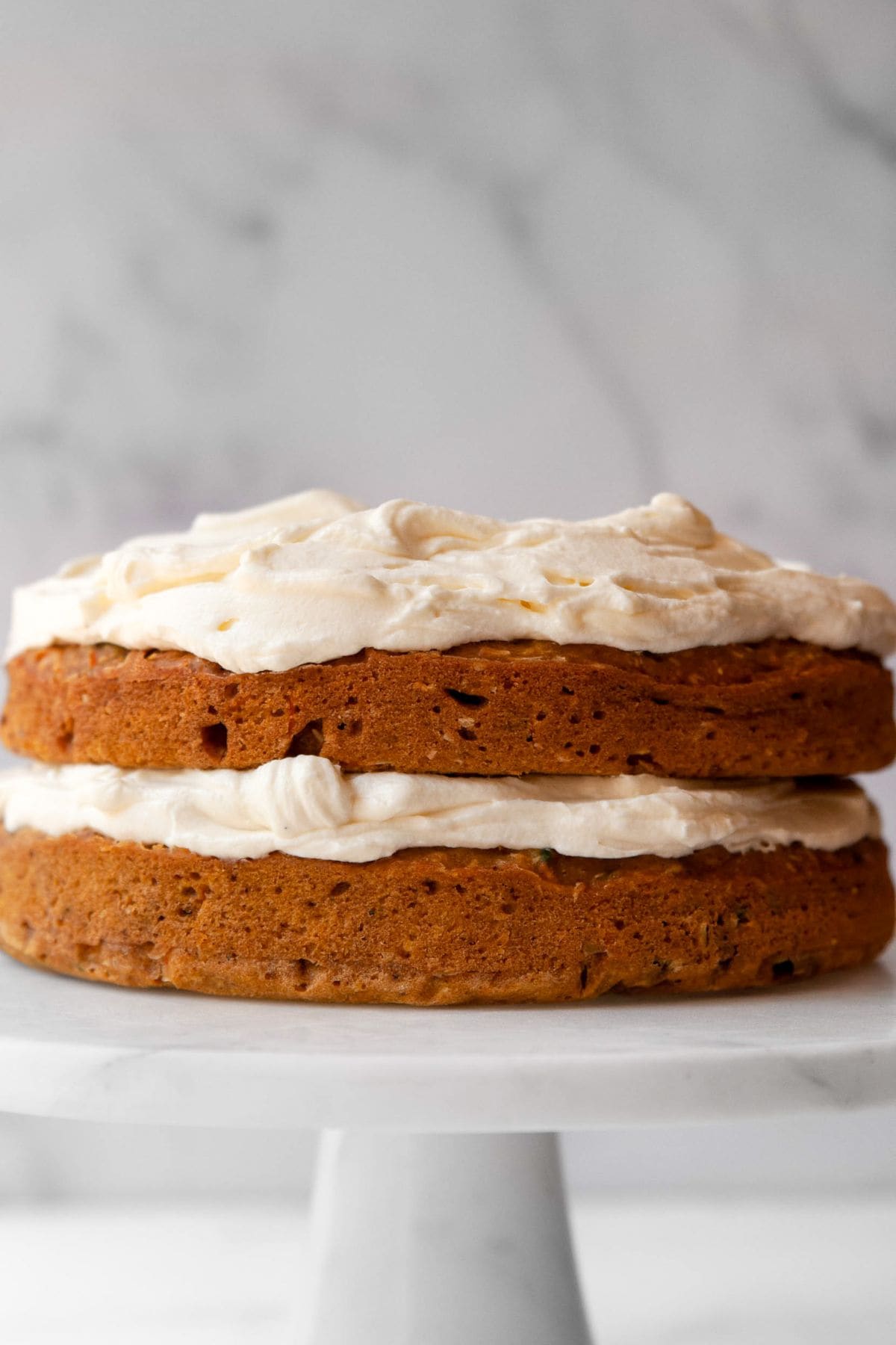 Two-layer pumpkin carrot cake with Greek yogurt cream cheese frosting between the layers and on top, sides left bare, on a white cake stand.