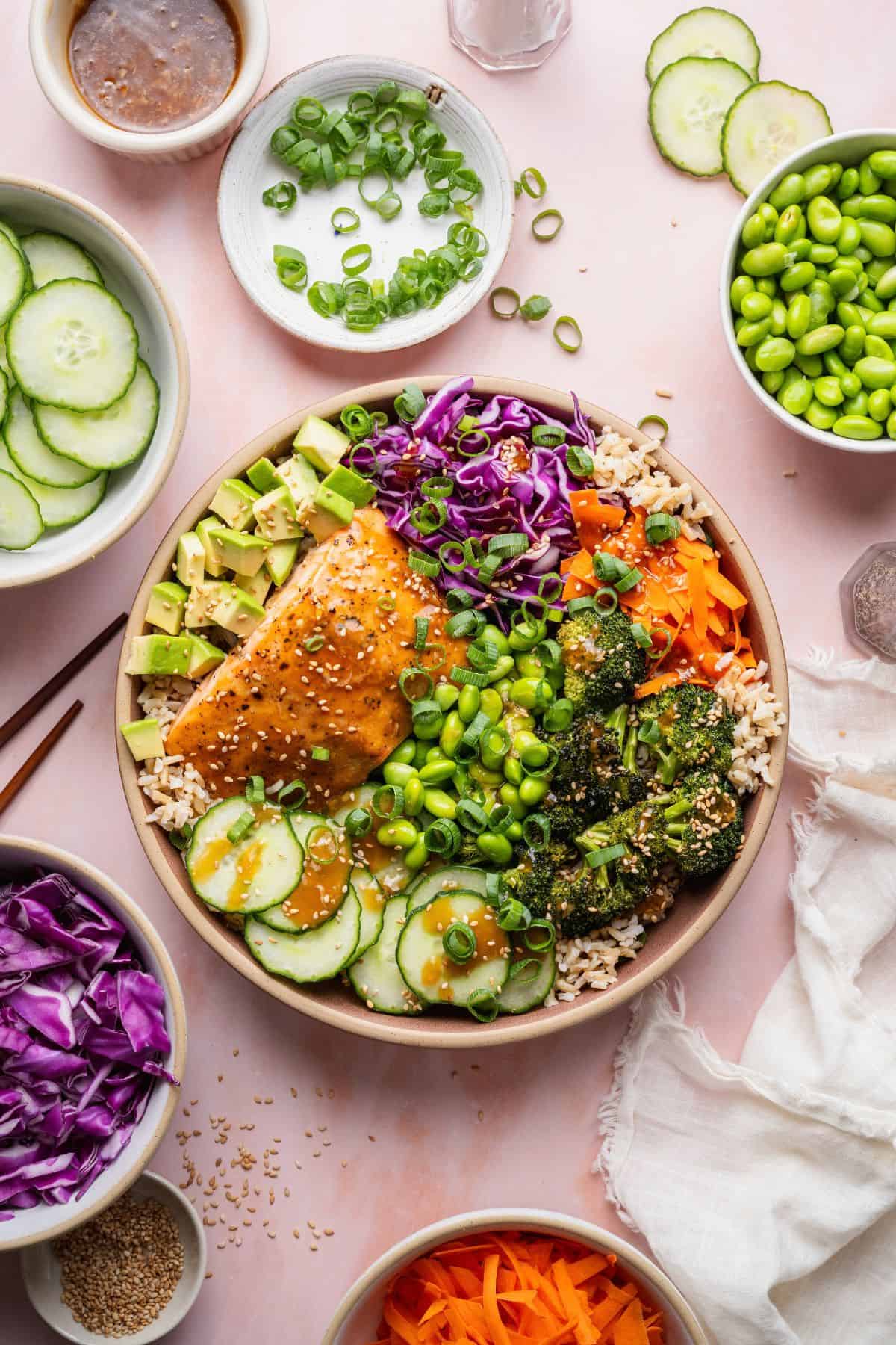 Teriyaki salmon bowl drizzled with homemade sauce, surrounded by cucumber, edamame, cabbage, and carrots.