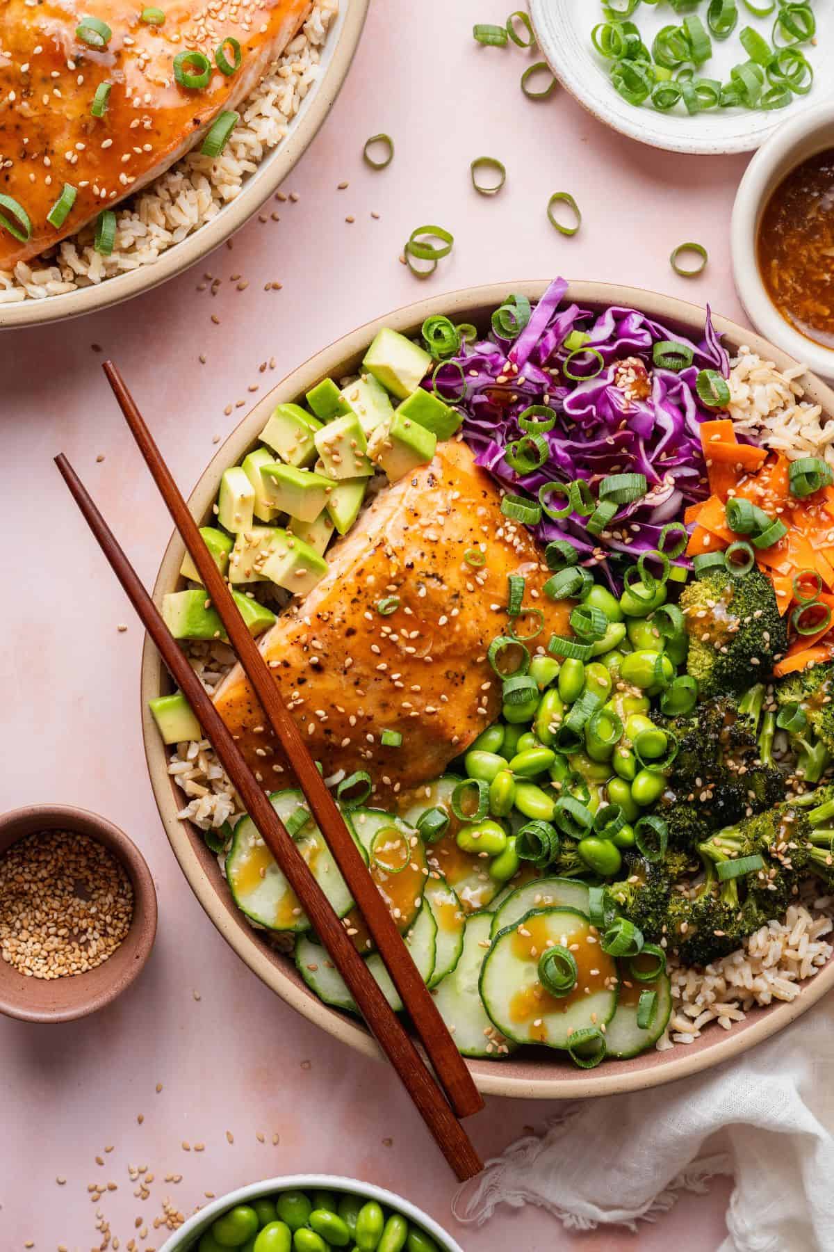 Teriyaki salmon with brown rice, roasted broccoli, edamame, and avocado in a bowl with chopsticks.