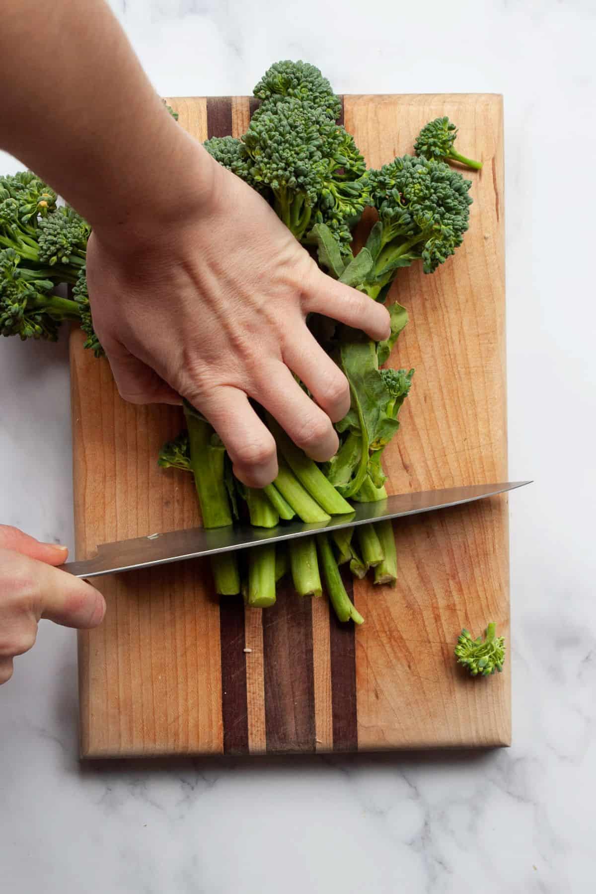 Trimming the ends off a bunch of broccolini with a knife on a wooden cutting board.
