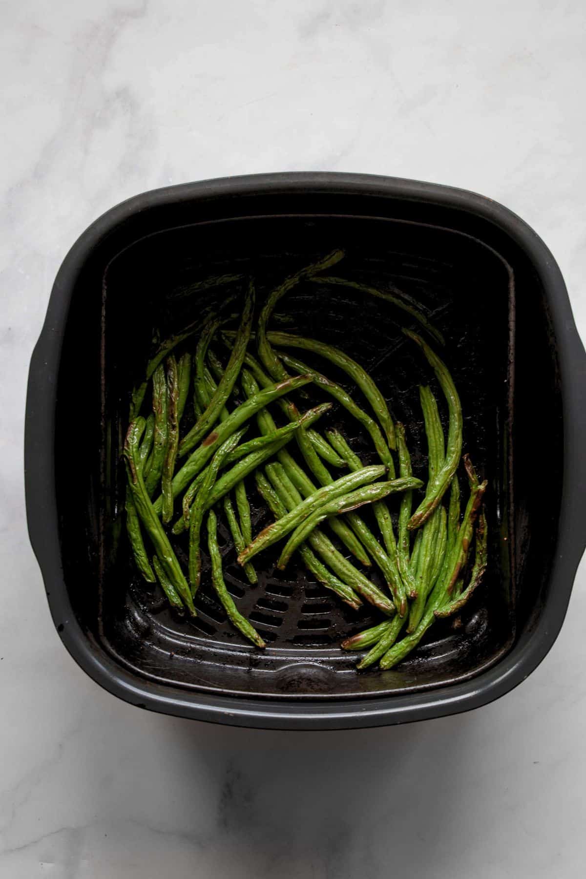 Seasoned raw green beans spread in an even layer in an air fryer basket.