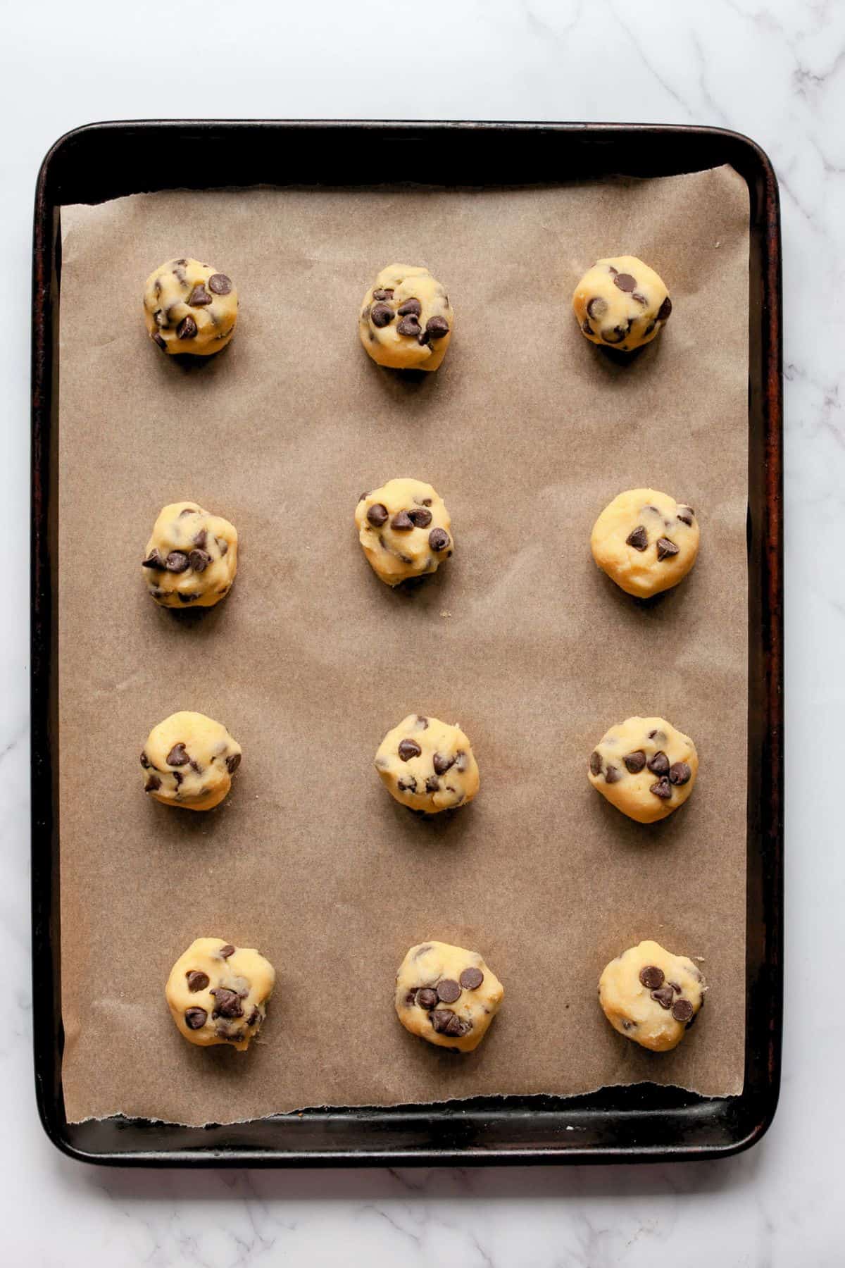 Twelve chocolate chip cookie dough balls on a parchment-lined baking sheet, ready to bake.