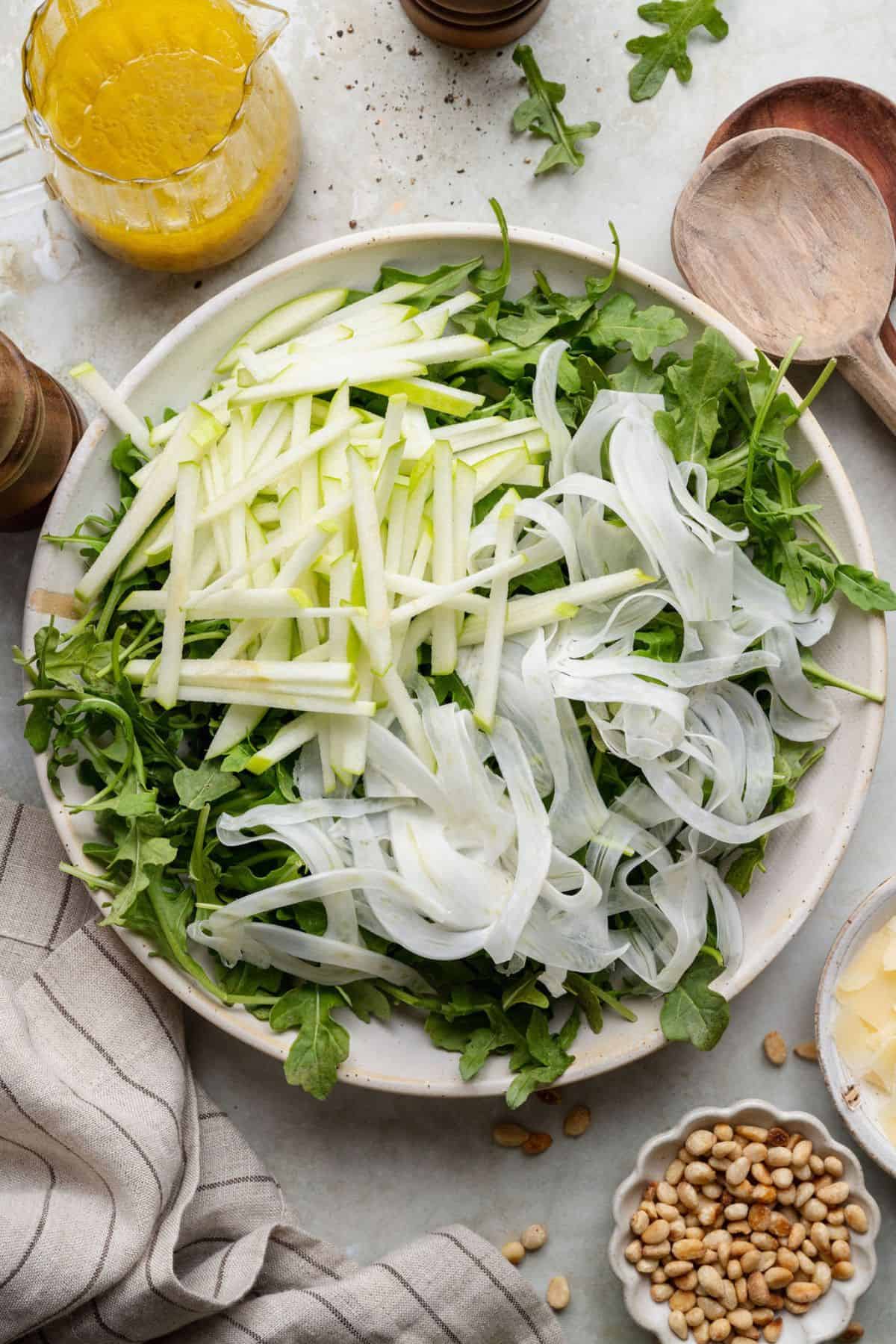 Assembling fennel arugula salad by layering shaved fennel and apple matchsticks over arugula before adding the dressing.