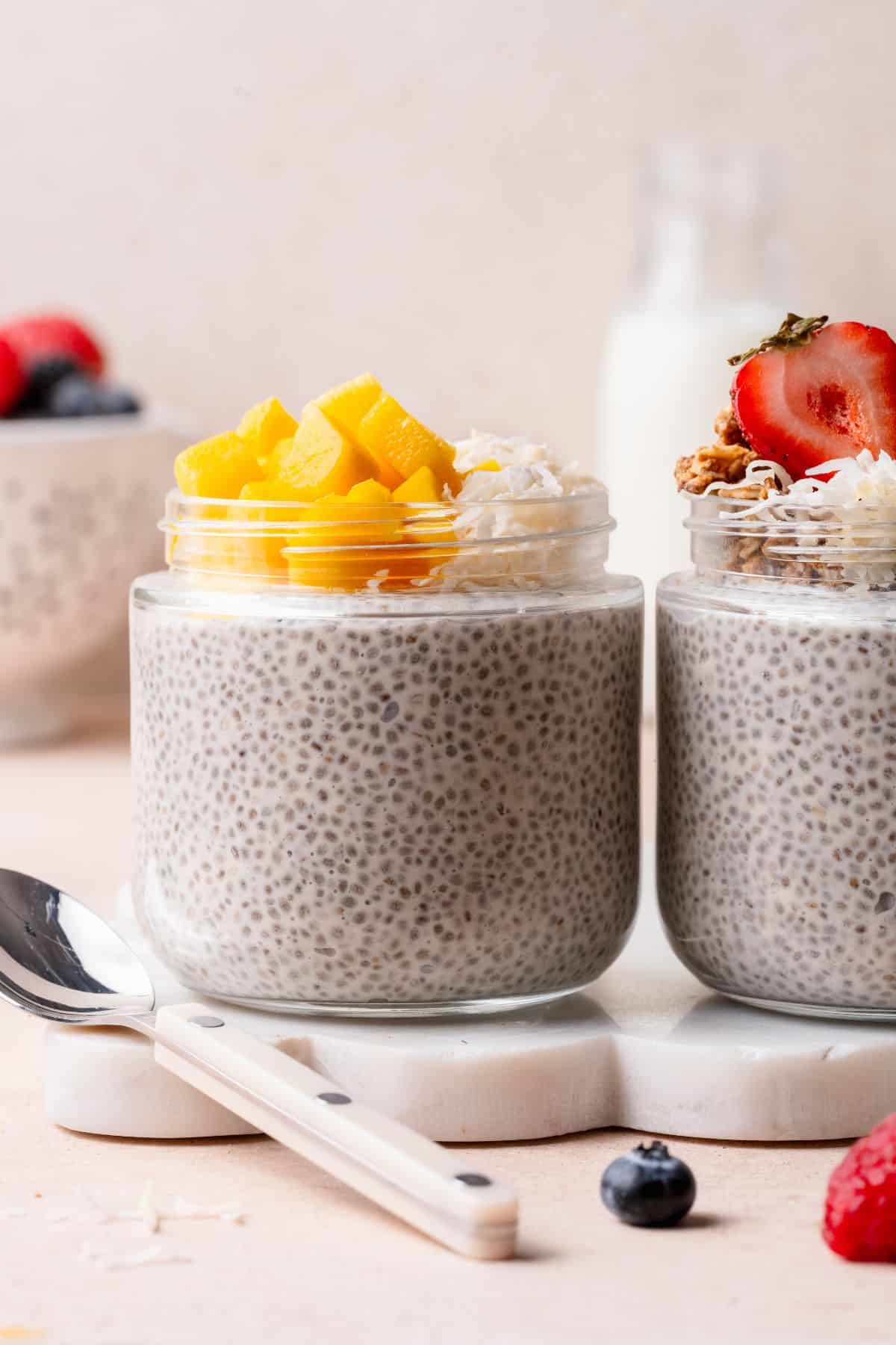 Two jars of coconut chia pudding, one topped with mango and one with strawberries and granola.