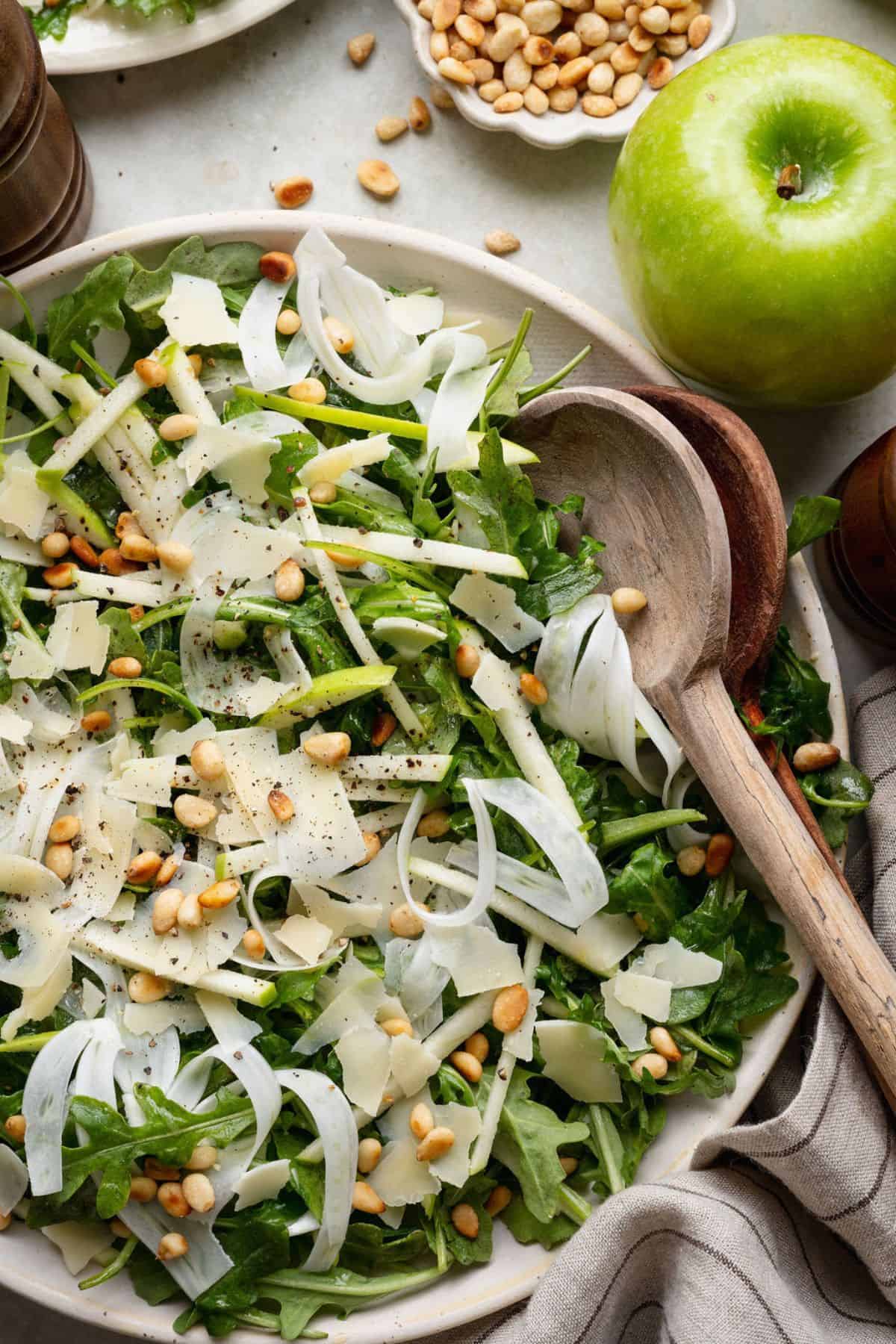 Fennel arugula salad with apple matchsticks and shaved Parmesan in a serving bowl with wooden spoons.
